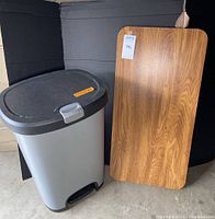 Vintage folding table with woodgrain Formica top standing beside a hefty plastic trash can with foot pedal lid, shown from front at slight angle on concrete floor with black backdrop.