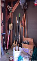 Various long handled garden tools including shovels, spades, rakes, broom, and short handled tools stored together with gloves and boxes.