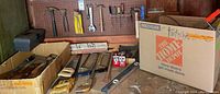 Wide view of various hand tools including multiple hand saws, hammers, screwdrivers, a lever, household oil, and a cardboard box on workbench.