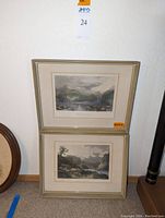 Two framed landscape prints stacked against a wall. Each has vintage-style nature scenes with mountains and water.