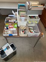 Table with assorted office supplies including note pads, pens, pencils, highlighters, index cards, staplers, paper clips, pushpins, metal rim tags, greeting cards, Skrip ink, magnifier, and hole punch in plastic drawer organizers.