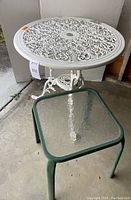 Photo showing white cast iron round patio table with peeling paint and detailed scrollwork top and legs, alongside small square green metal frame glass top side table.