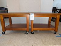Pair of small brown wood tables with rectangular tops and lower shelves, each on four black casters wheels for mobility. Tables measure approximately 20x14x15 inches.
