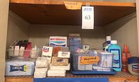 Shelf with multiple unopened first aid and toiletry items including boxes of sterile pads, bars of soap, Epsom salt, towelettes, bottles of lotion and mouthwash, and plastic bins holding toiletries.