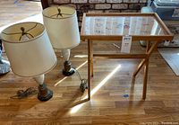 Photo showing two decorative table lamps with white porcelain bodies, brass bases, off-white shades, and a wooden TV tray with floral-patterned glass top.
