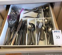 Top view of flatware organized in a drawer tray, showing a variety of knives, forks, spoons, and serving utensils including some with white handles and others polished stainless steel, also includes a sealed plastic bag with additional flatware.