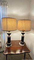 Pair of vintage table lamps shown on wooden side table with shades on. Beige/tan fabric shades with brown trim. Lamps turn on and emit warm light.
