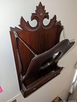 Wall mounted wood storage unit with carved ornamental top, hinged angled door featuring an oval mirror and carved leaf/branch accents, metal chains on sides supporting the door open, and small holes at bottom likely for keys or small items