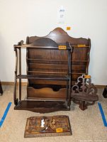 Photo showing three different wooden shelves arranged together on the floor against a wall along with an intricate carved wooden bookend and a separate carved wooden panel.