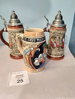 Front view of all three beer steins, two with metal lids and one without lid, showing detailed carved relief and decorative elements.