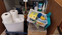 Overview of various cleaning supplies arranged in bins under a sink including paper towels, sponges, cleaning cloths, spray bottles, and plastic bins.