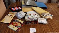 Photo showing the overall lot on a table, including three albums/binders (black Ambassador Album, blue binder, USA album), a tin, envelopes, and plastic bags with stamps.