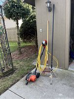 Black and Decker electric edger alongside yellow extension cord, metal garden rake, and tree trimmer with long handle, positioned outdoors next to a shed.