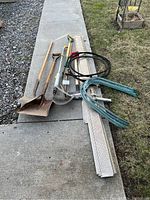 Overall photo showing three long-handled shovels, a black coiled small garden hose with spray nozzle, several perforated metal gutter guard pieces, and green metal garden stakes laid on sidewalk and nearby grass.