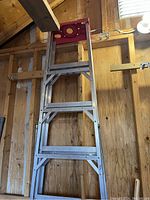 Tall aluminum ladder with red plastic top leaning against wooden wall in shed.