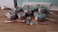 Full set view of six stainless steel Lagostina pots with lids and three kitchen utensils on floor