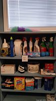 Shelf displaying multiple bottles of cleaning sprays, detergent boxes, blue dryer balls, fabric softener sheets, steel wool pads, clothes pins and duster