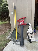 Photo showing a red Toro electric leaf blower with black blower tube and beige leaf collection bag hanging on blower. Next to blower are two garden rakes: one green plastic leaf rake with wooden handle, and one smaller metal landscaping rake with yellow fiberglass handle.