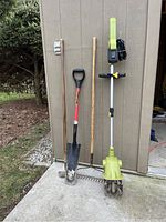 Full view of rototiller and garden tools including shovel, rake, hoe, and digging fork against shed wall