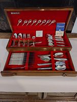 Full view of silverplate flatware set in wooden box with red velvet lining including spoons, forks, and knives arranged systematically in compartments.
