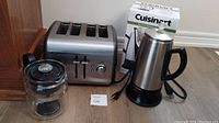 Photo of the Cuisinart kettle, KitchenAid 4-slice toaster, and Gemco heat-resistant glass pot. Items placed on floor against wall, showing details and condition of each appliance.