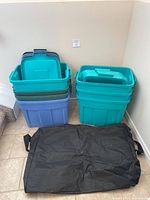 Seven Rubbermaid storage bins stacked with lids to the side next to black duffel bag on tiled floor