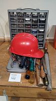 Tools arranged on a wooden surface showing the hard hat, hand tools including wooden-handled files and screwdriver handles, and a multi-compartment organizer case for screws and nails.