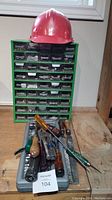 Wide photo showing red hard hat on top of green multi-drawer organizer with hand tools arranged on gray socket set case