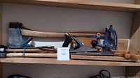 Wide shot of shelf displaying various hand tools including axes, saws, hammer, and hand planer.