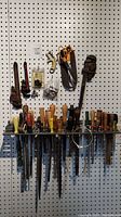 Overview of assorted hand tools displayed on a pegboard and hanging tool holder: multiple chisels, screwdrivers, metal files, hanging holder, scissors, and pipe wrenches.