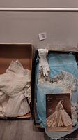 Box containing vintage white wedding dress folded with lace and satin fabric visible, white bridal gloves, pearl beaded bridal headpiece, and promotional photo/card of wedding dress