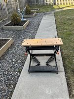 Front view of the Black & Decker Workmate workbench on a concrete pathway outdoors with gravel and garden beds in the background.
