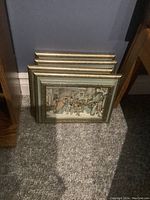 Four gold-toned framed prints stacked against wall, showing backs and edges, confirming four items in the lot.