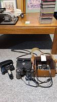 Collection of cameras, binoculars, camera cases, and flash arranged on carpeted floor under wooden table.