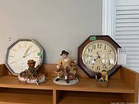 Two quartz clocks placed on a wooden shelf alongside three figurines: a bear in a canoe, a man on a bench, and a small fishing bear figurine.