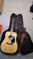 Full view of acoustic guitar inside open black soft shell case with brown strap