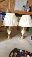 Pair of metal base table lamps with white pleated shades standing on garage floor with various storage items around. Both lamps have pull chains and matching designs.