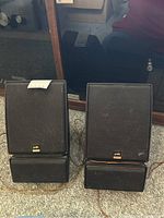Pair of black Polk Audio Monitor Series 2 speakers with cords and removable fabric grilles, placed upright on the floor showing front sides.