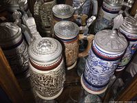 Collection of multiple German beer steins on shelf showing various painted and relief designs with pewter lids.
