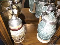 Four ceramic beer steins with metal lids arranged on wooden surface near mirrored background showing reflections and details