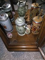 Three beer steins including a gray textured one, a green waterfowl hunting-themed one, and a colorful human figure-themed stein with pewter lids on wooden shelf.