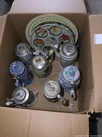 Top view of all beer steins and metal trays inside a cardboard box showing variety and some damage