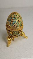 Front and side angle view of the antique enamel gilt egg on the gold-tone three-legged stand, showing blue and green enamel with gold filigree detail.