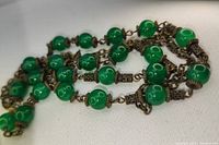 Close-up of green round beads linked with ornate antique brass-toned metal caps and intricate chain elements, showing bead and metal detail.