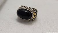 Front and side views of a large vintage ring featuring a polished black onyx stone set in a silver band with intricate scrollwork and gold-tone heart details.