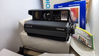 Photo of black Polaroid Spectra 2 instant camera with flash and lens, resting on a white surface near blue original box and manuals.