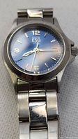 Front view of ESQ wristwatch showing blue dial with Arabic numerals and date window, stainless steel case and band.