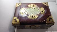 Front angled view of the vintage jewelry box showing dark wood, central jade panel, and brass corner and border decorations.