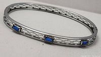 Oval silver-tone metal bracelet featuring blue rectangular stones and geometric cutout filigree