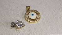 Close-up photo of a circular evil eye pendant featuring a gold-tone frame with clear stones and a central white and blue eye design, placed on a textured fabric background.